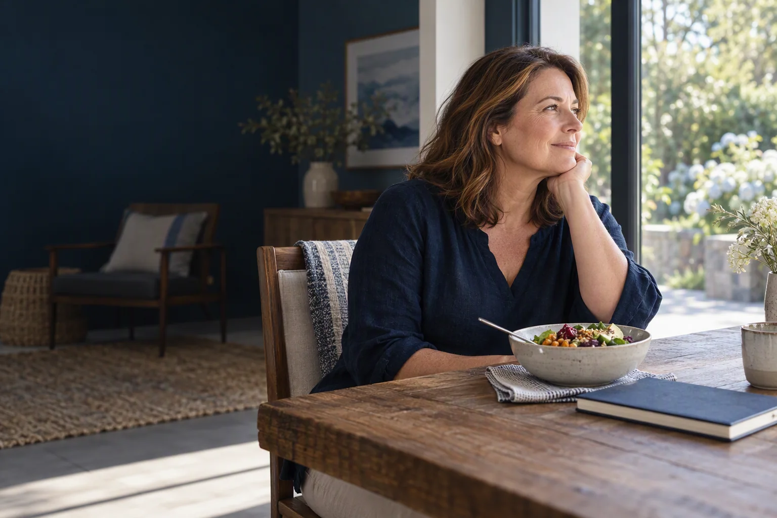 A woman in a bright home, with a meal and notebook on a table.